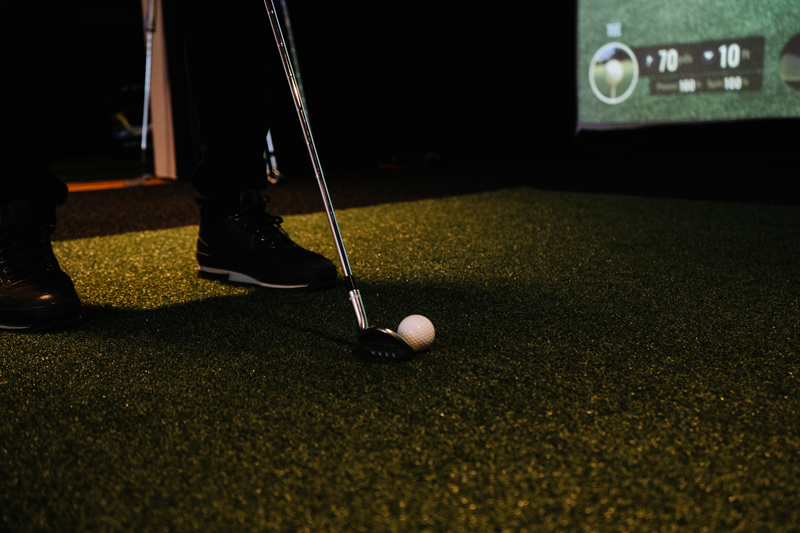 A person stands on artificial green turf, holding a golf club next to a golf ball, with Golf Simulators displaying scores on the virtual screen in the background.