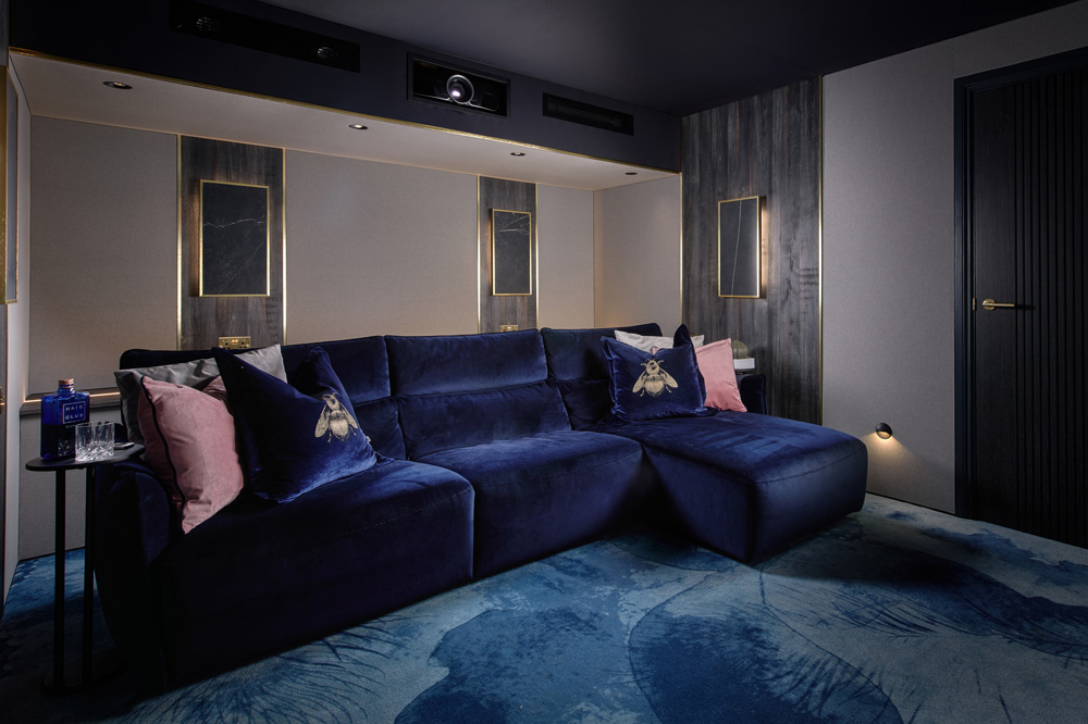 A modern home cinema room with a dark blue velvet sectional sofa, decorative pillows, a blue patterned rug, side table with drinks, and dim wall lighting, creating a cosy and luxurious atmosphere.