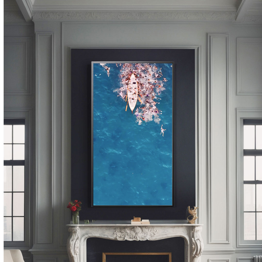 A large framed photo of a boat surrounded by swimmers in blue water hangs above a white marble fireplace in a bright, elegantly decorated room with tall windows and white paneled walls.