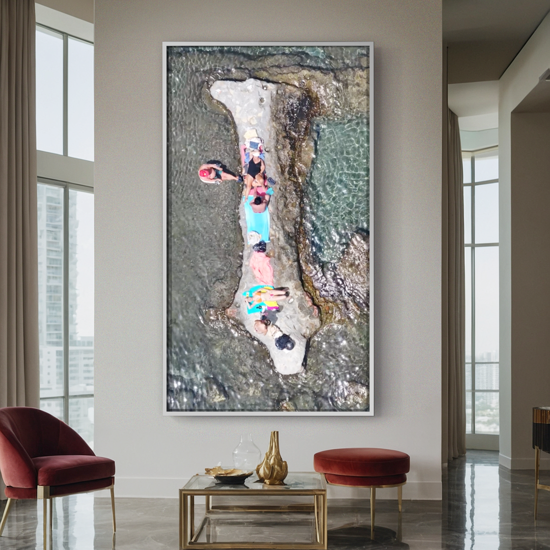 A large framed aerial photo of people lying on a rock surrounded by water is displayed on a wall in a modern living room with tall windows, red chairs, and a gold-accented coffee table.