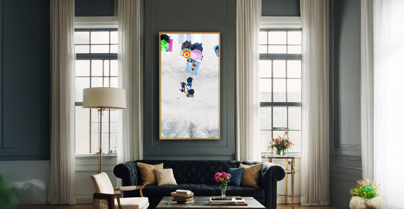 A stylish living room with tall windows, white curtains, and a dark sofa features a large, vertical framed photo from a Digital Art Gallery of people in winter clothing walking in snow, hanging above the sofa.