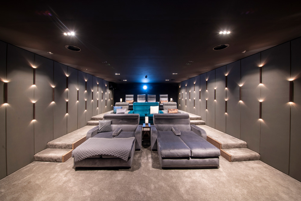 Modern home cinema with tiered grey seating, including luxurious lounge chairs and sofas, soft carpeting, and ambient wall lighting, creating a cosy and upscale atmosphere. A blue light glows from the projection area at the back.