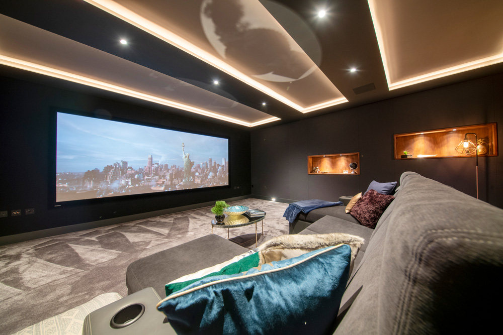 A modern home cinema room with a large projection screen showing a cityscape, grey sectional sofa with colourful cushions, decorative shelves, and recessed ceiling lights. The space feels cosy and luxurious.