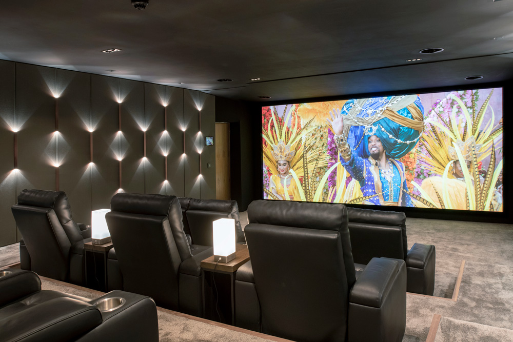 A modern home theater with black leather recliners, small side tables with lamps, and a large screen displaying a colorful parade scene. The room has wall lighting and gray carpet.