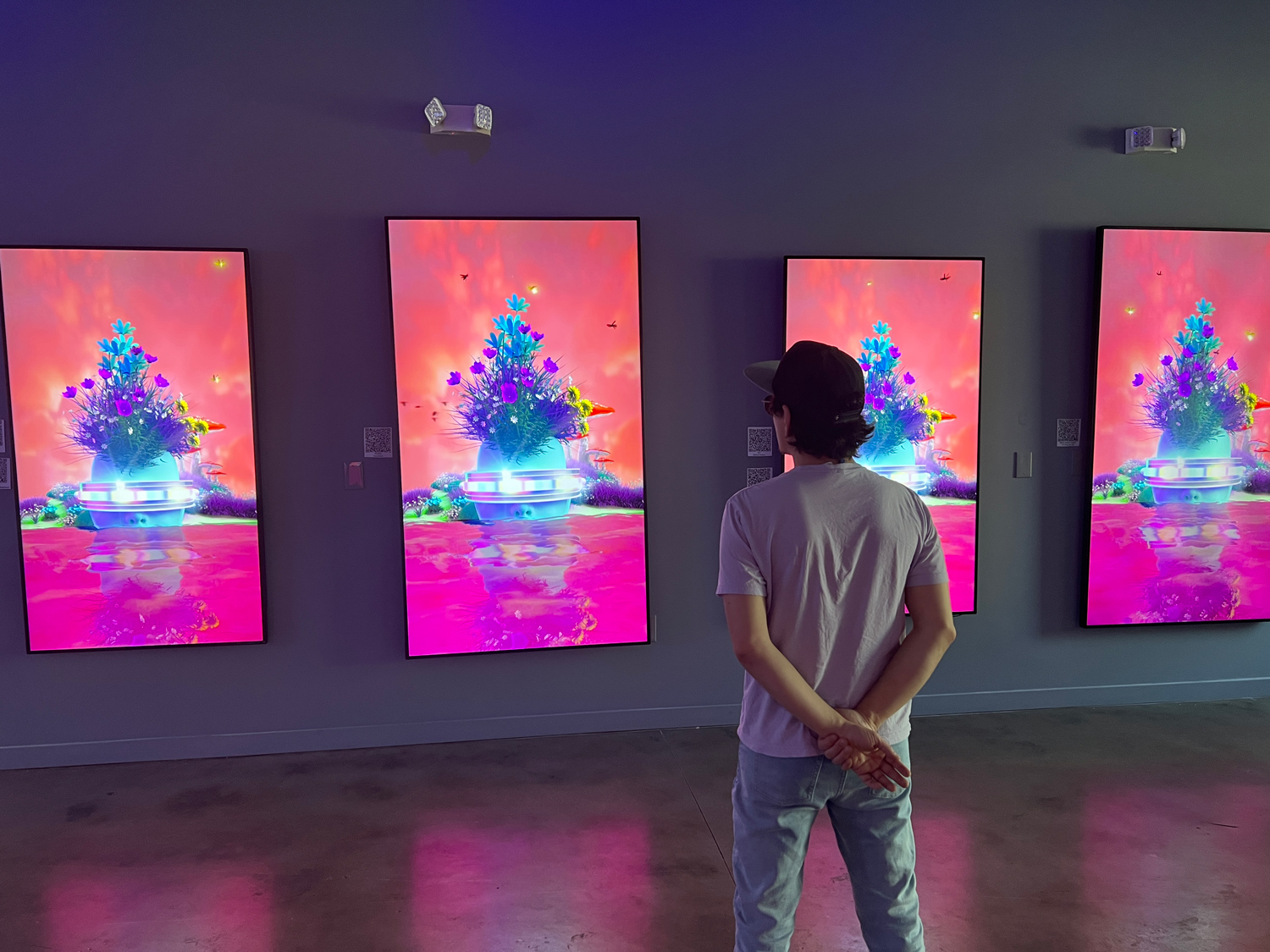 A person stands with hands behind their back, observing three vibrant digital screens displaying colorful, futuristic landscapes with glowing trees and pink skies in an art gallery setting.