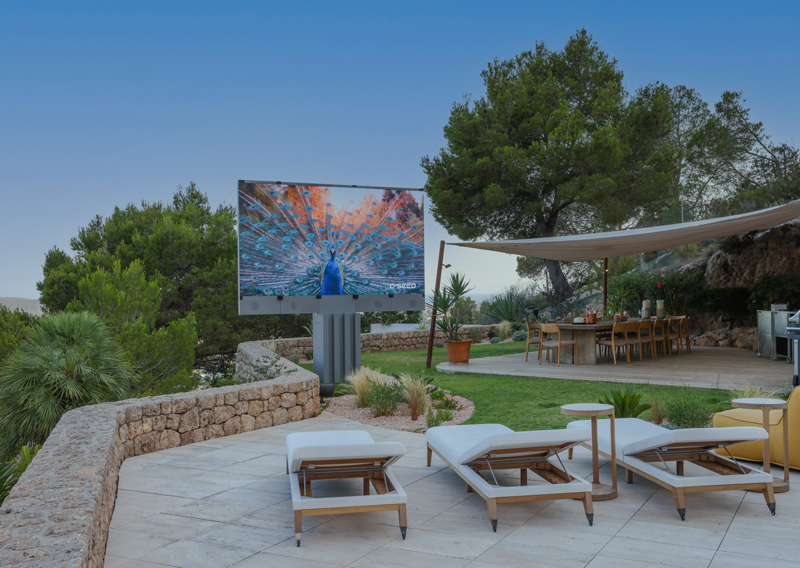 Outdoor patio designed for outdoor entertainment, with several lounge chairs facing a large screen displaying a peacock image, surrounded by greenery, stone walls, a shaded dining area, and trees in the background under a clear sky.