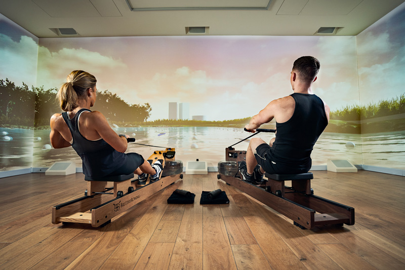 Two people in athletic gear use indoor rowing machines in front of a large screen displaying a virtual river scene, creating an immersive rowing experience in a modern, well-lit Wellness Room.