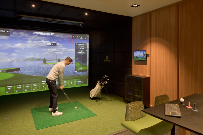 A person enjoys golf simulators in a modern indoor room, standing on a green mat and swinging a club toward a screen displaying a virtual golf course. Golf clubs and seating are visible in the space.