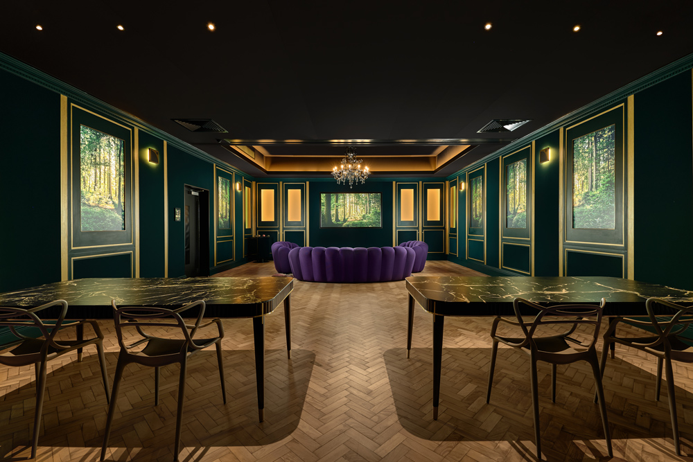 A luxurious Home Nightclub room with dark green walls, gold trim, a chandelier, parquet flooring, tables with chairs, a curved purple sofa, and framed nature art on the walls.