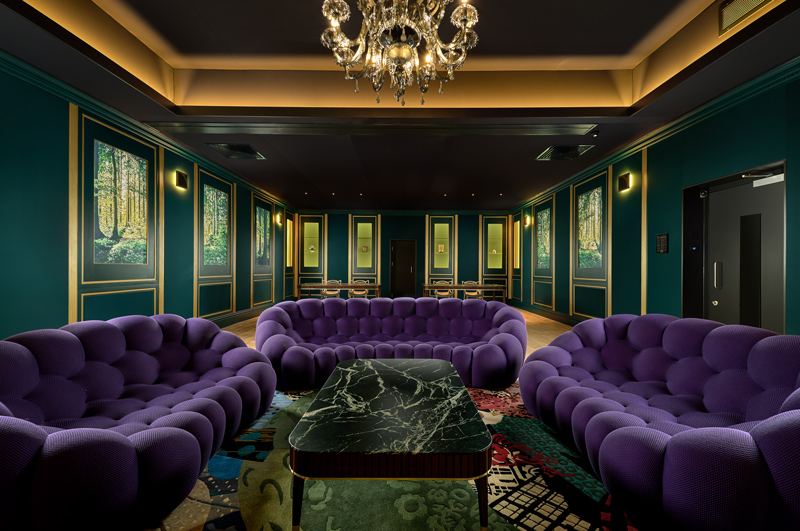 A stylish lounge with deep green walls, purple tufted sofas, a black marble coffee table, and a chandelier evokes an upscale home nightclub vibe. Forest-themed wall art, a patterned rug, and gold trim accents complete the luxurious space.