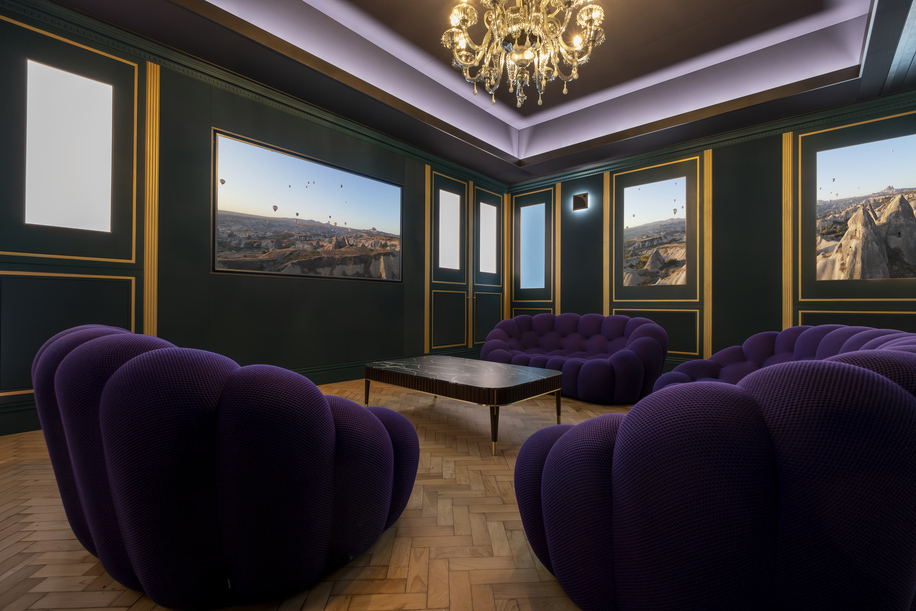 A stylish lounge with dark green walls, large landscape paintings, and a chandelier evokes a chic home nightclub vibe, featuring plush purple sofas arranged around a central coffee table on a herringbone wood floor.