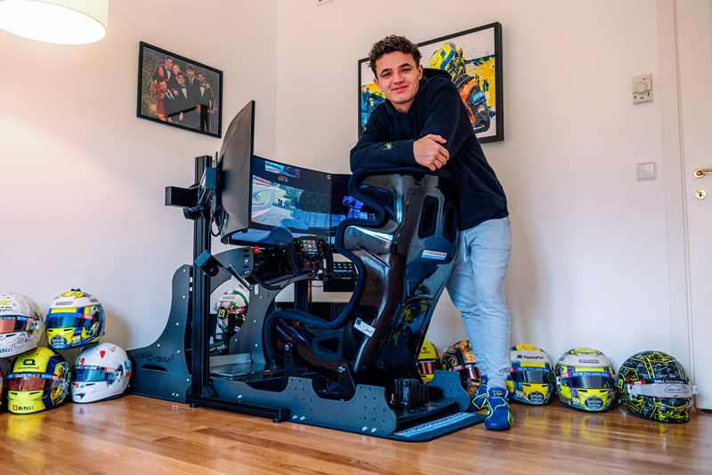 Lando Norris stands smiling next to a Cool Performance driving simulator setup in a room decorated with racing helmets on the floor and motorsport-themed artwork on the walls.