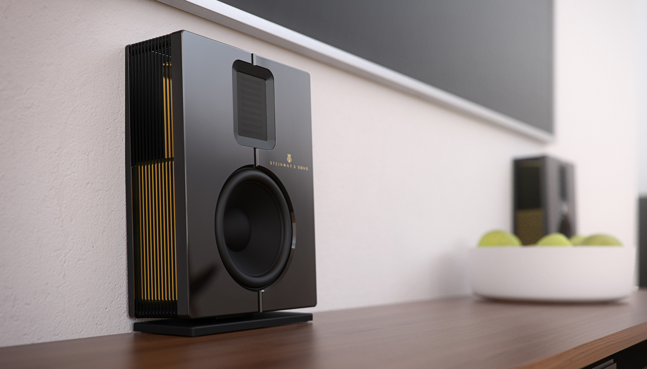 A sleek black Steinway Lyngdorf speaker with gold accents sits on a wooden surface near a white bowl of green apples, with another identical speaker in the background against a white wall.