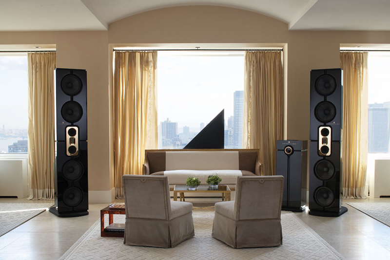 A modern living room, designed for exceptional listening rooms, features two tall black speakers beside a sofa, two beige armchairs, a coffee table, and large windows with gold curtains overlooking a cityscape.