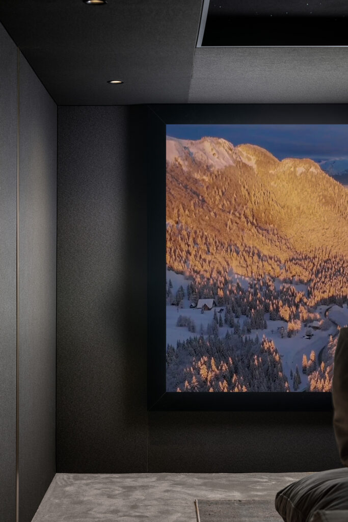 A modern, dark-walled private cinema room with a large screen displaying a scenic, sunlit mountain landscape covered in snow. Part of a persons shoulder is visible in the lower right corner.