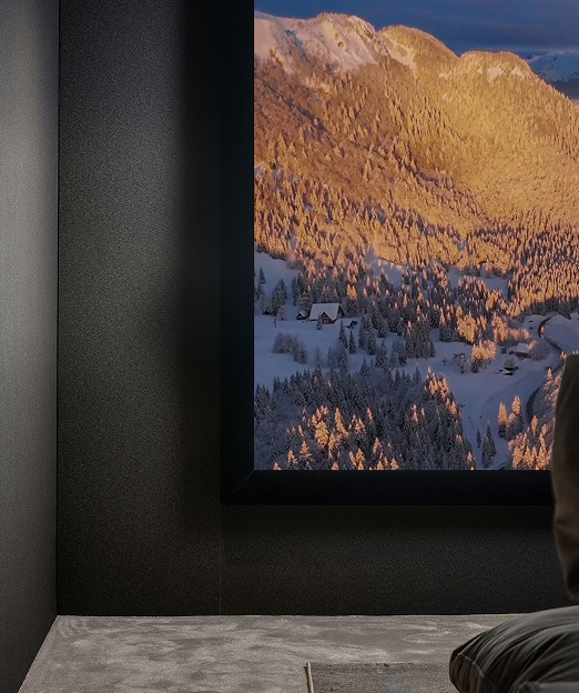 A dark, modern room with a large wall-mounted screen showing a snowy mountain landscape lit by golden sunlight, featuring trees and a small cabin among the snow.