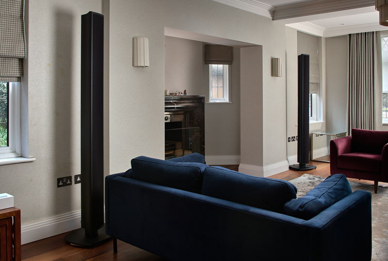 A modern living room designed for Listening Rooms, featuring a navy blue sofa, two tall black speakers, wood flooring, a fireplace, and large windows with patterned blinds. Light walls and maroon armchairs complete the cozy, stylish space.