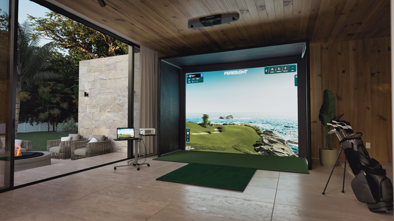 A modern indoor Golf Simulators setup in a wooden room, featuring a large screen displaying a golf course, an artificial turf mat, golf clubs, and an open sliding door leading to a patio with outdoor seating.