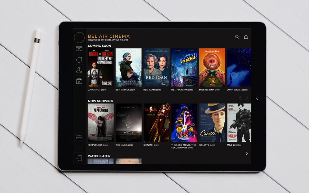 A tablet displays a streaming app called Bel Air Cinema showing movie posters and titles in sections like Coming Soon, Now Showing, and Watch Later, with a stylus beside it on a white wooden surface.