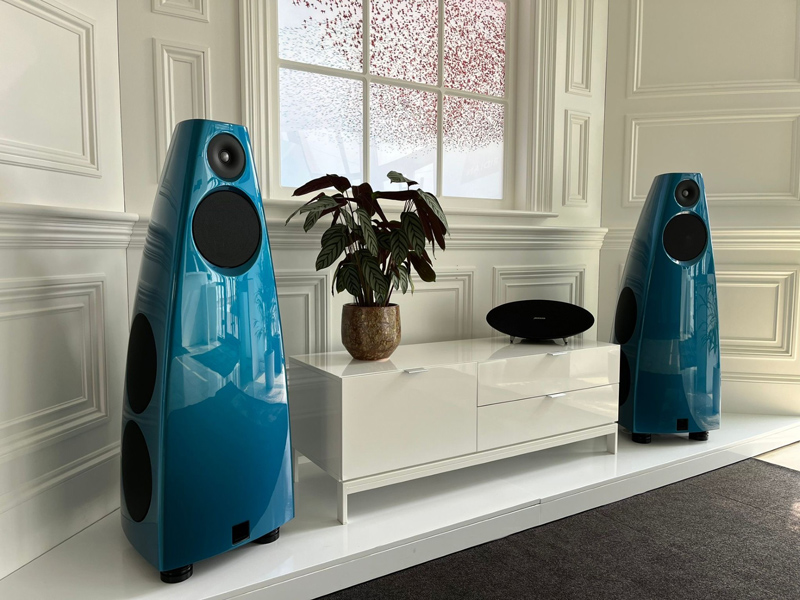 Two large, sleek blue speakers stand on either side of a white cabinet with a potted plant and a black speaker on top, creating an inviting listening room against a white-paneled wall with a window in the background.